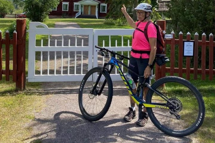 Person på cykel utanför en vit grind och rött staket. I bakgrunden är en Hälsingegård. 