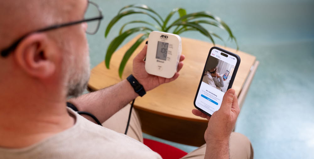 En person sitter ner och mäter sitt blodtryck med en digital blodtrycksmätare. Personen håller mätaren i ena handen och en mobiltelefon i den andra, där en app med vårdrelaterat innehåll visas. 