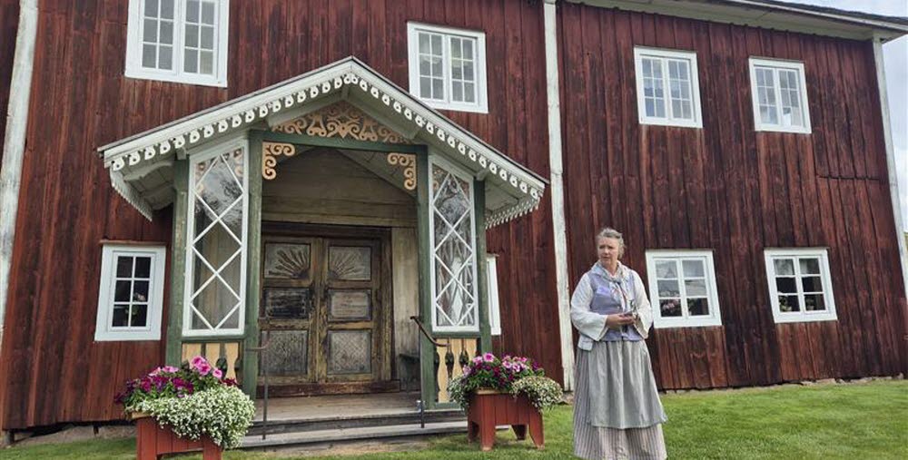 En person i folkdräkt står framför en stor rödmålad träbyggnad med snickarglädje vid entrén.
