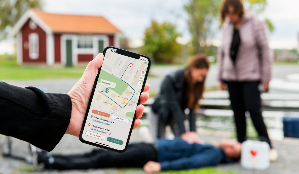 Hand med mobiltelefon, och i telefonen bild på en karta. I bakgrunden en suddig bild på tre personer, där hjärt-lungräddning (HLR) pågår. Bilden är arrangerad och en kvinna står vid sidan, en annan ligger på marken. En tredje ger HLR. 